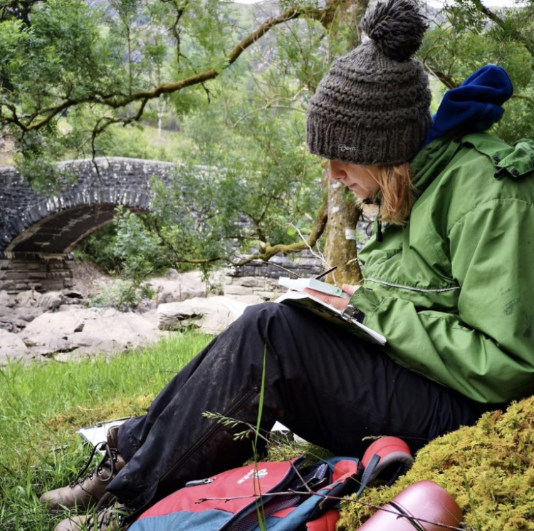 A woman in a bobble hat sits on a river bank, sketching